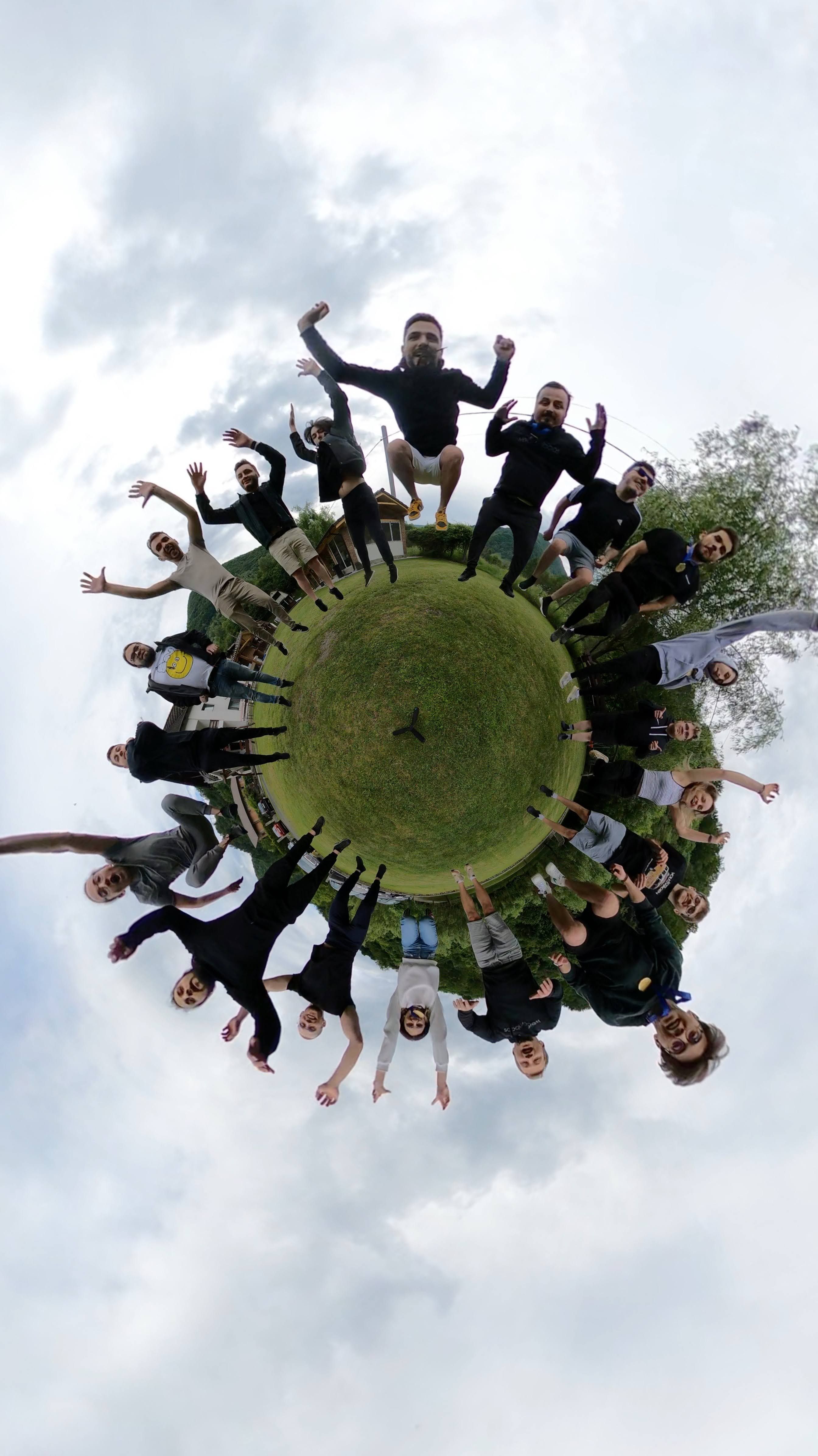 A group of people jumping in the air outdoors captured in a 360-degree “tiny planet” style photo with grass and cloudy sky surrounding them.