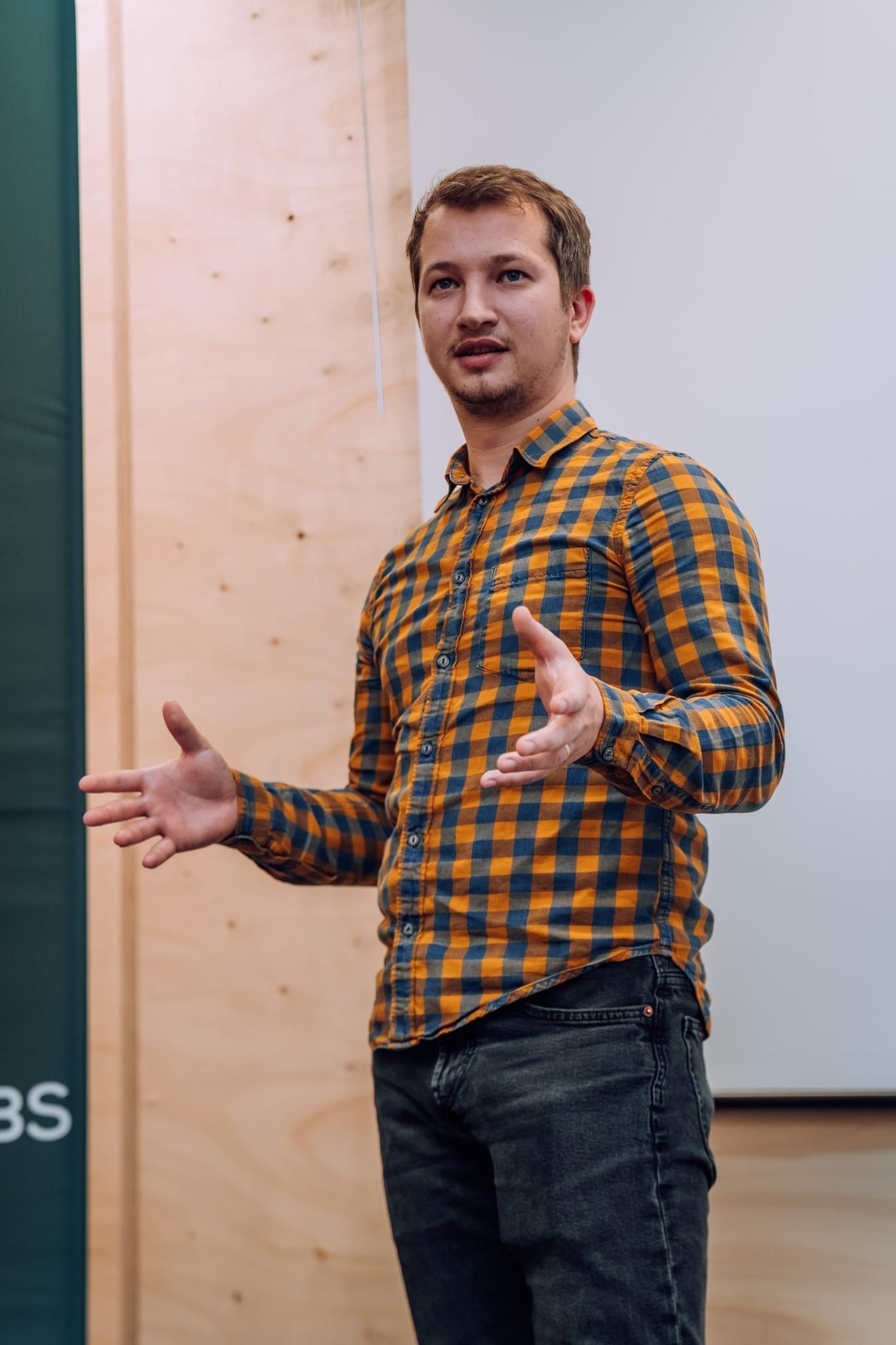A man in a plaid shirt giving a talk while gesturing with his hands during a presentation in a conference room.