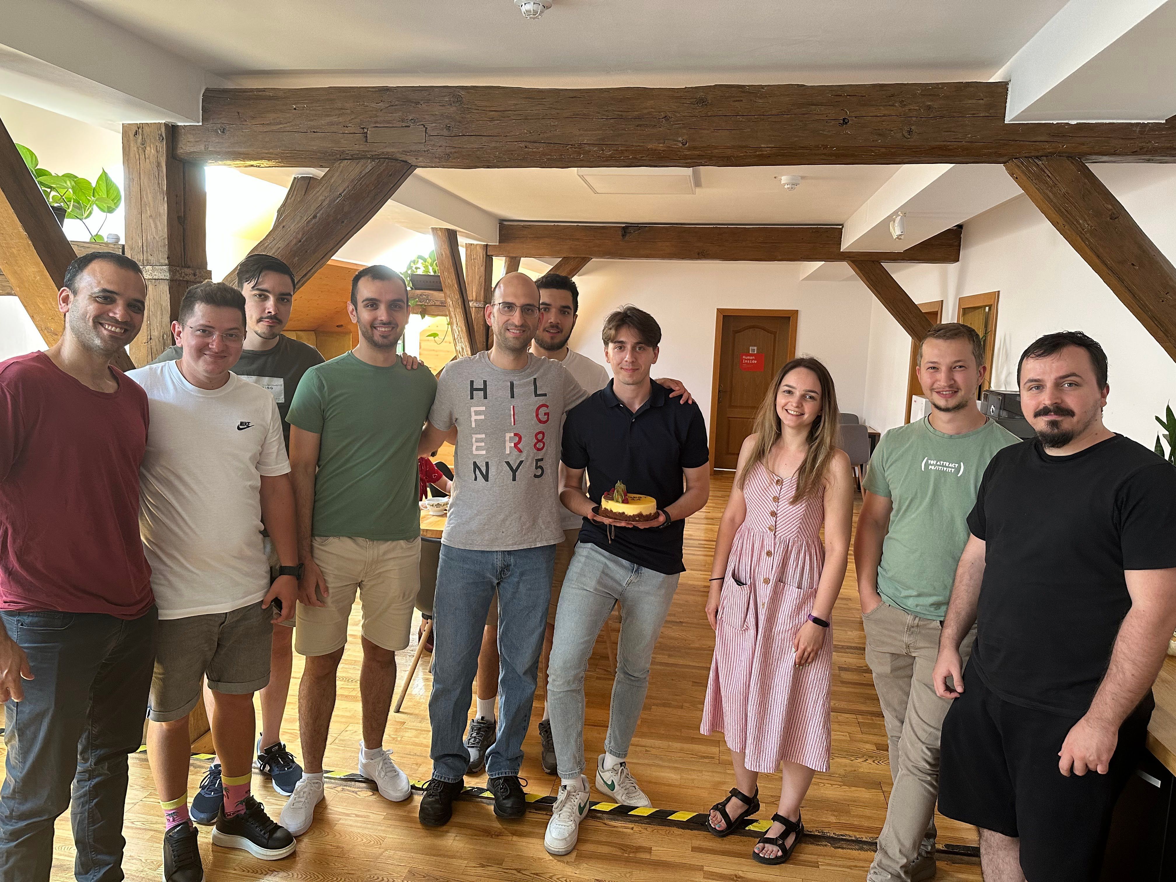 A group of coworkers standing together indoors under wooden ceiling beams while one person holds a small birthday cake, smiling for a team photo.