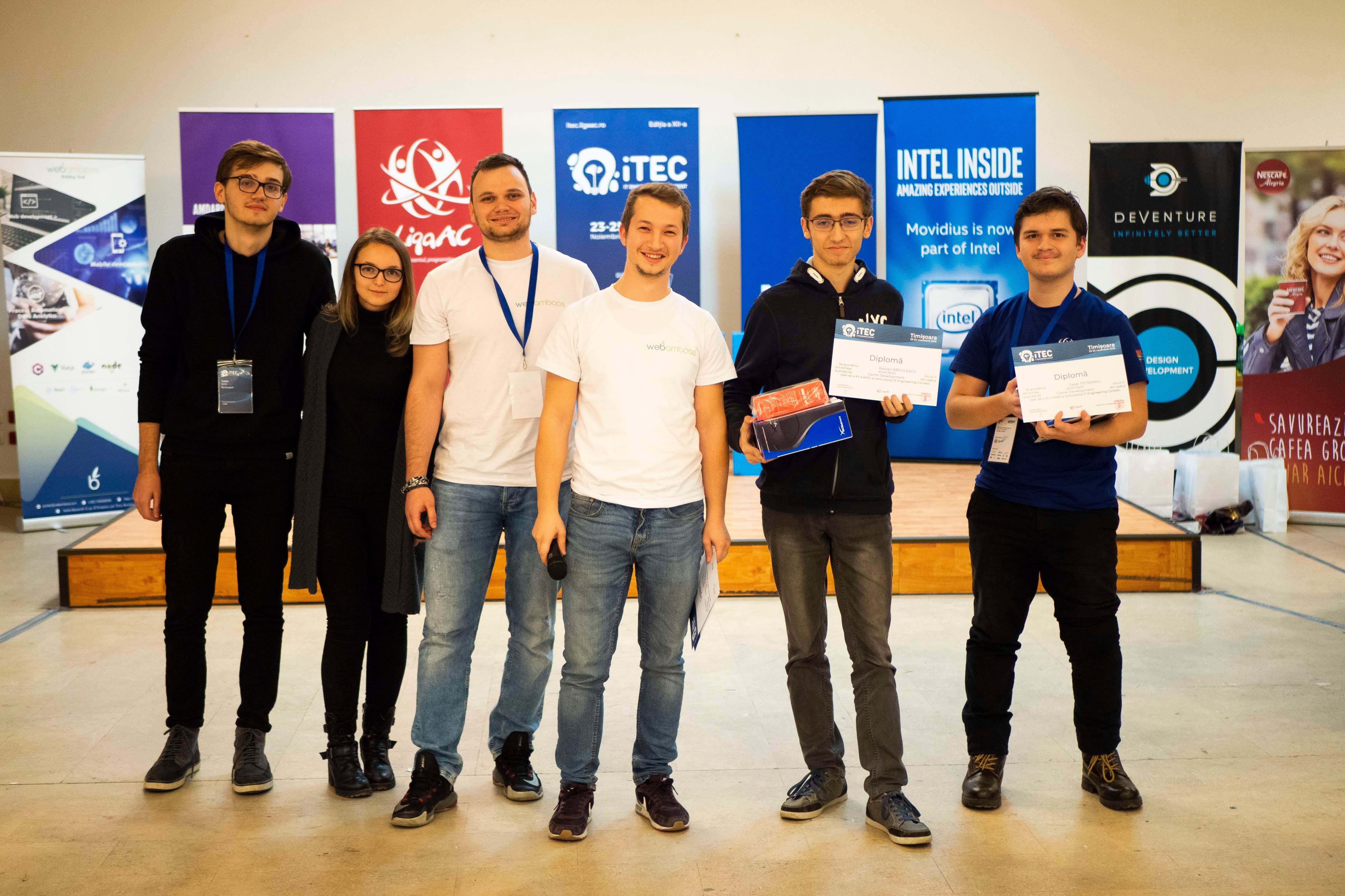 Six young adults standing on a stage in front of event sponsor banners, two holding diplomas and a boxed prize during an iTEC competition ceremony.